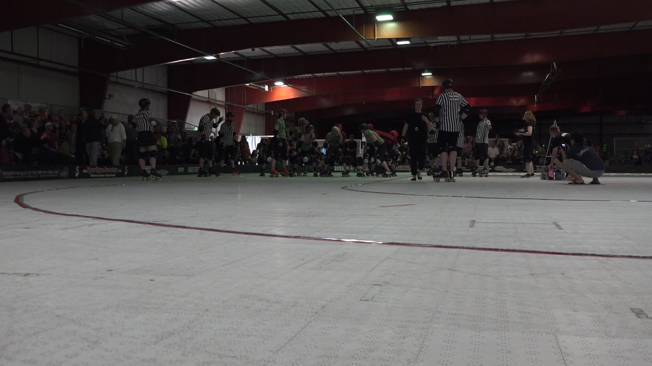 Low view across an indoor skating rink with players, officials, and spectators near the boards.