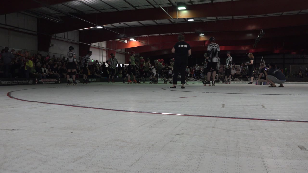 Low view across an indoor skating rink toward referees, skaters, and spectators.