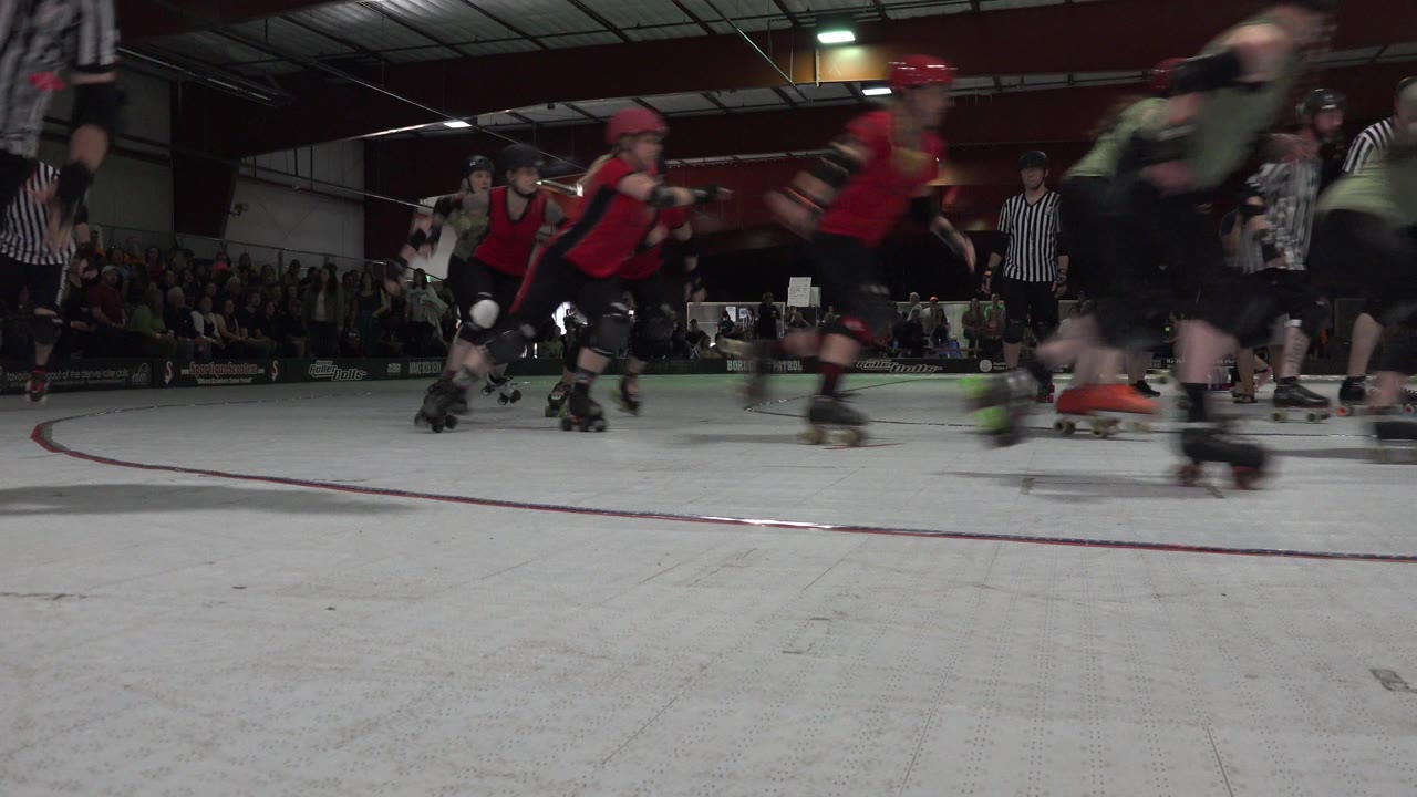 Low-angle Denver, Colorado frame of roller derby skaters in helmets and pads moving across an indoor rink with spectators and referees behind them.