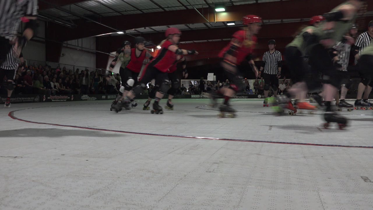 Denver, Colorado roller derby action continues with blurred skaters in red and dark uniforms passing a low camera as referees and a seated crowd remain in the background.
