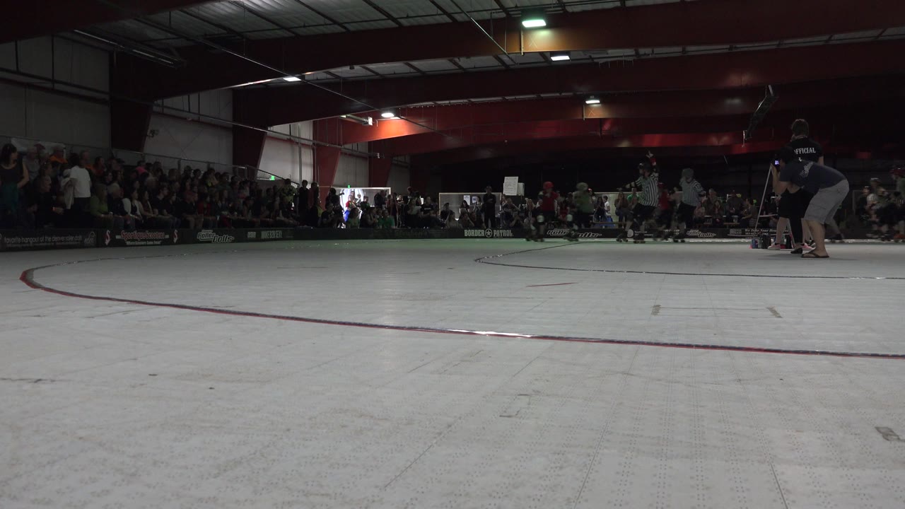 At 0:00 in Denver, Colorado, the low camera looks across a pale roller rink floor toward distant skaters, striped referees, and a seated crowd under dark indoor lighting.