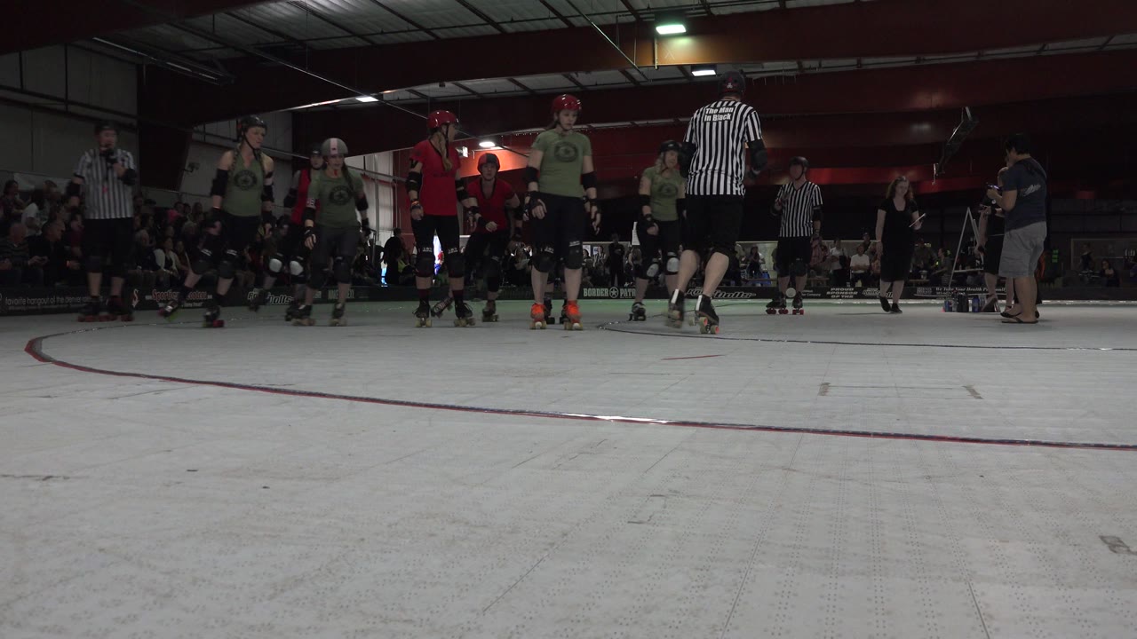 Skaters and referees on an indoor rink with spectators behind them.