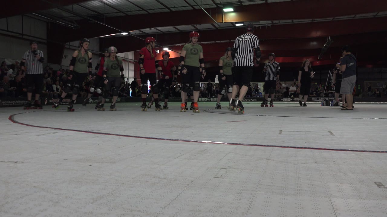 Denver, Colorado rink view with skaters in red and green tops near the center line, referees on roller skates, and a seated crowd along the back wall.