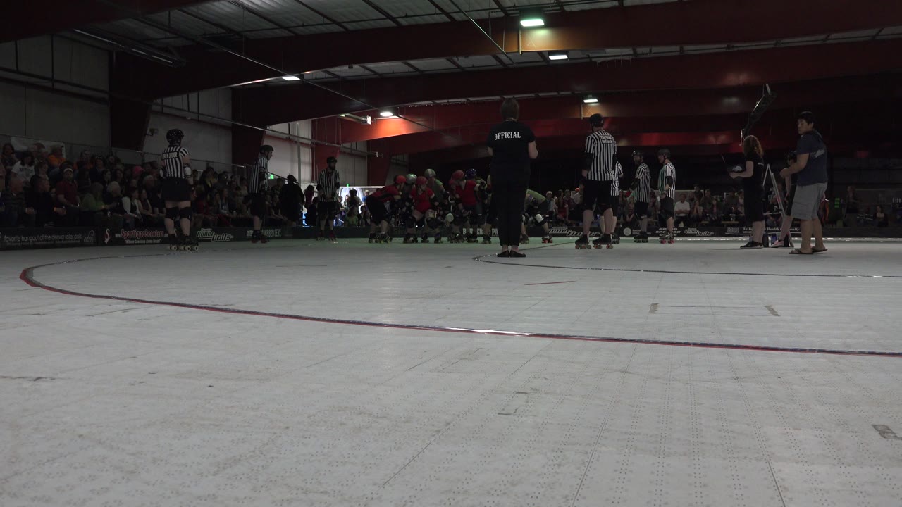 At 0:01, the Denver rink remains framed from floor level, with a striped referee near center and a cluster of skaters and spectators behind the track boundary.