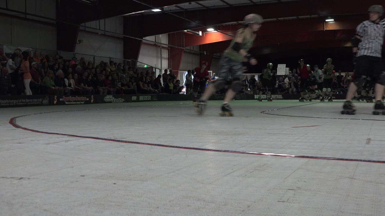In Denver, Colorado, a helmeted roller skater passes near a striped referee on an indoor rink while spectators sit in bleachers behind them.