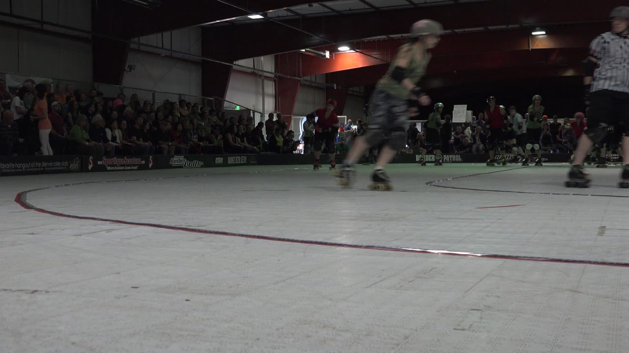 A skater blurs through the foreground of the Denver indoor rink while a referee stands to the right and the seated crowd fills the background.