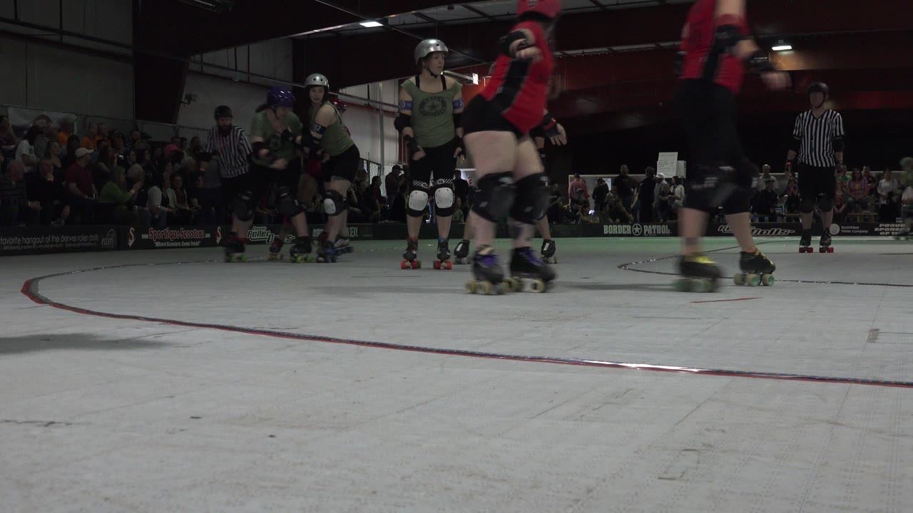 Roller derby skaters and a referee on an indoor rink with spectators behind them.