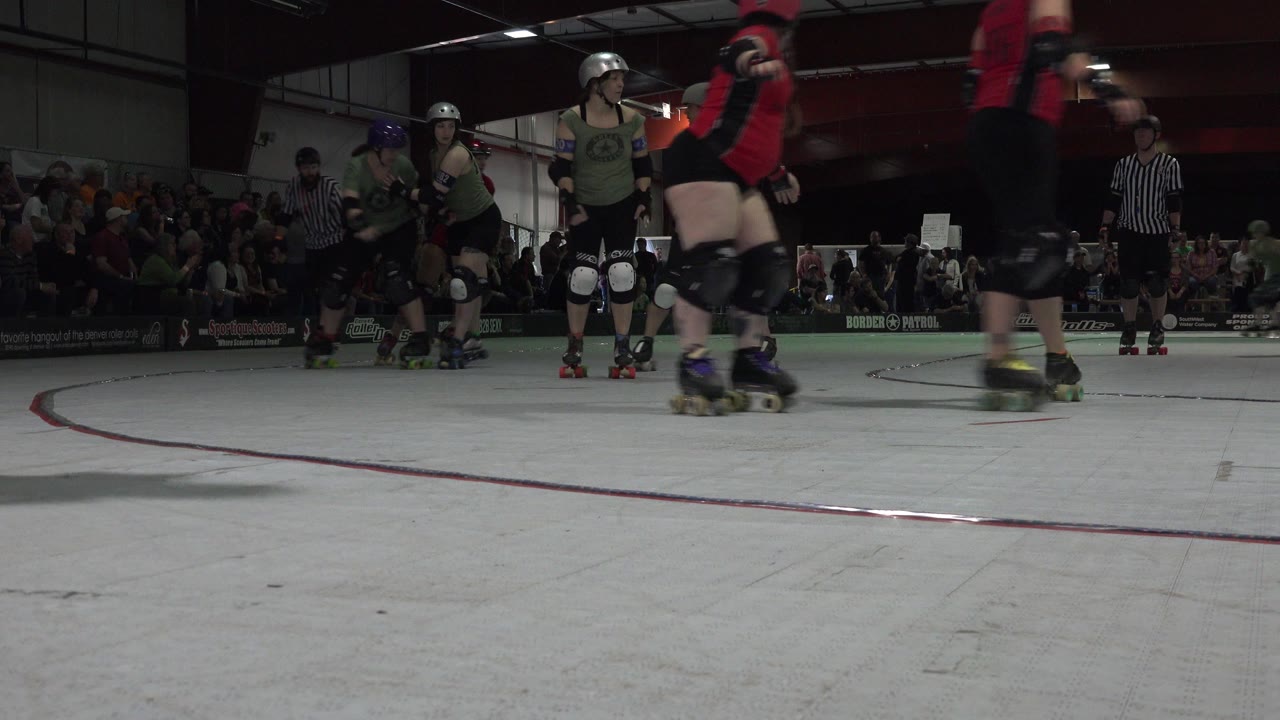 Roller derby players in green and red gear skate past a low ground camera in Denver, Colorado, with the crowd and referees visible beyond the rink boundary.