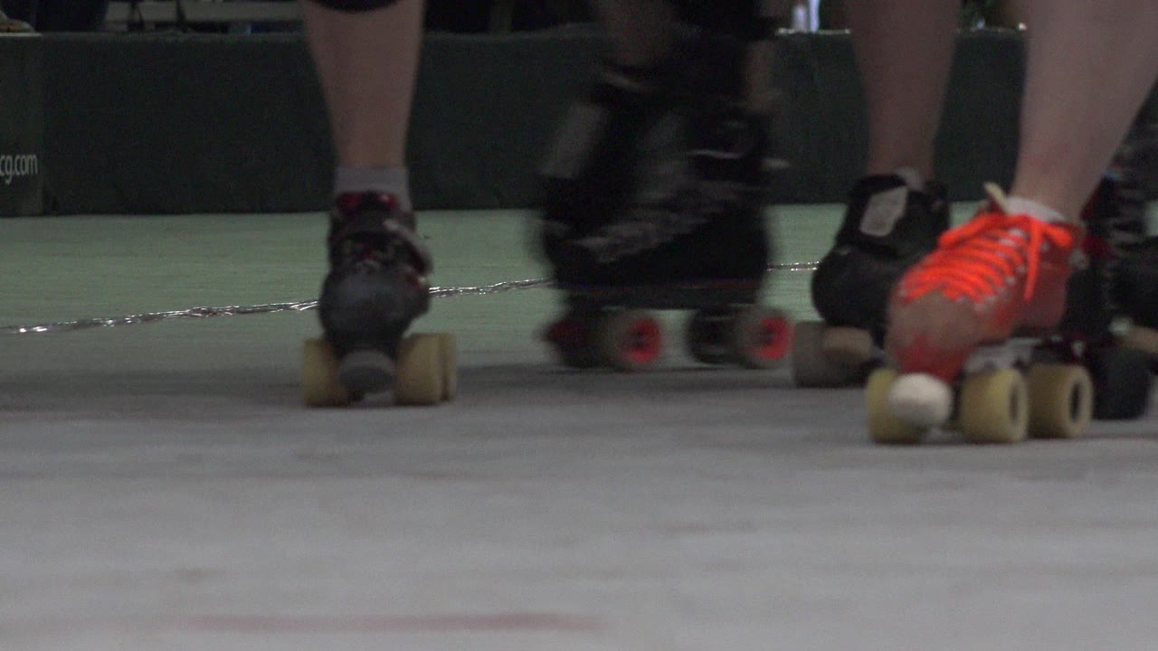 Floor-level Denver rink view with black and orange roller skates clustered in the middle distance and a blurred skate passing near the lens.