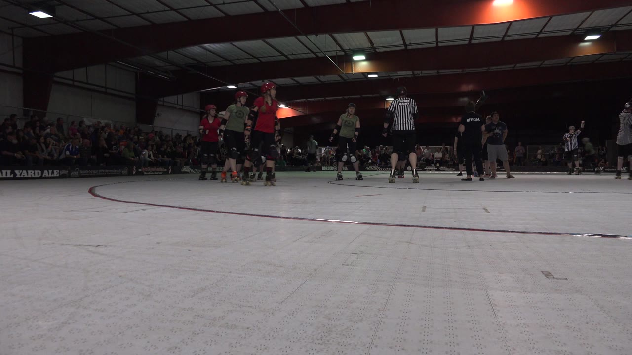 Helmeted skaters and referees on an indoor rink with spectators behind them.