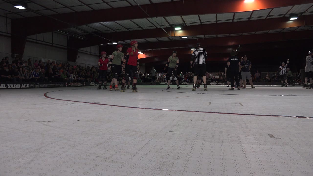 At 0:01 in Denver, Colorado, skaters in red and green tops remain near the center of the indoor rink with referees and a crowd behind them.