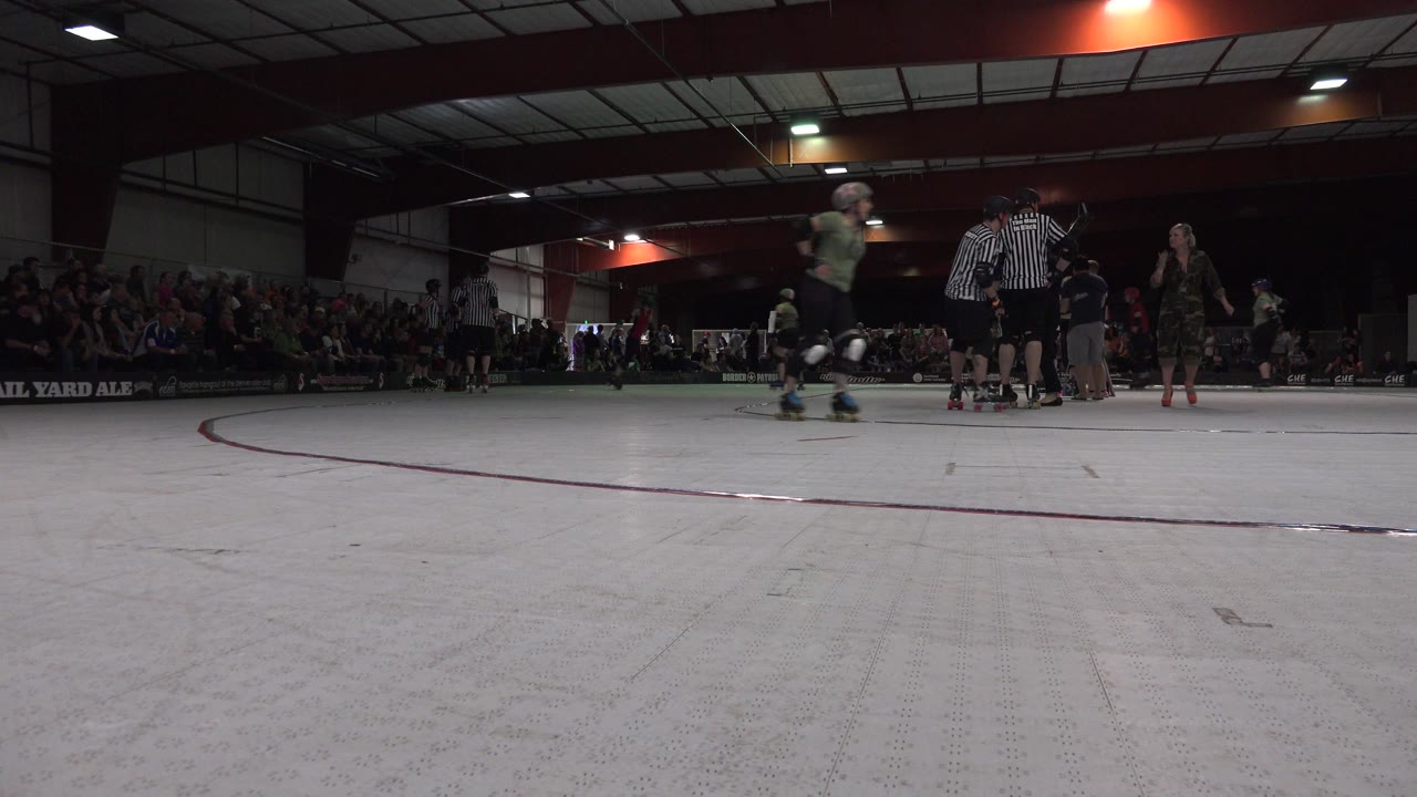 Denver indoor rink frame with a skater crossing the left side, referees near center, and a seated crowd stretching across the back wall.