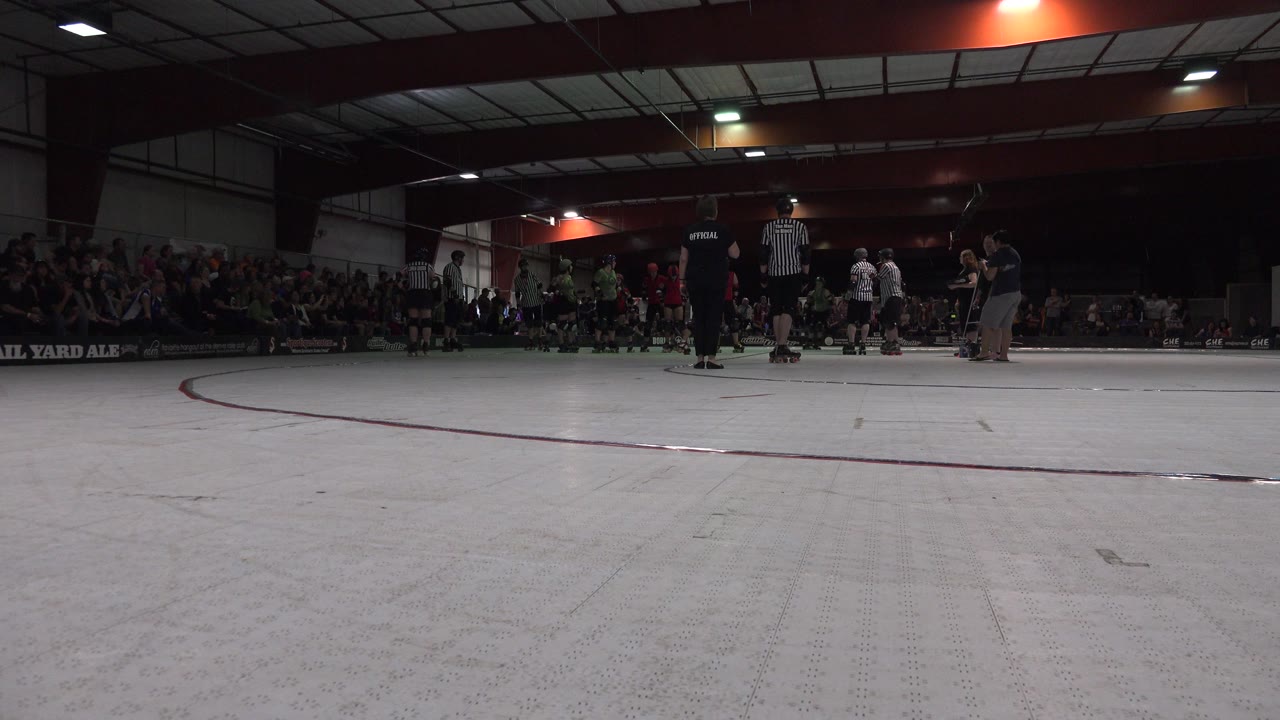 Low rink-side frame in Denver, Colorado shows the pale track foreground with roller derby skaters, striped referees, and seated spectators across the far side.