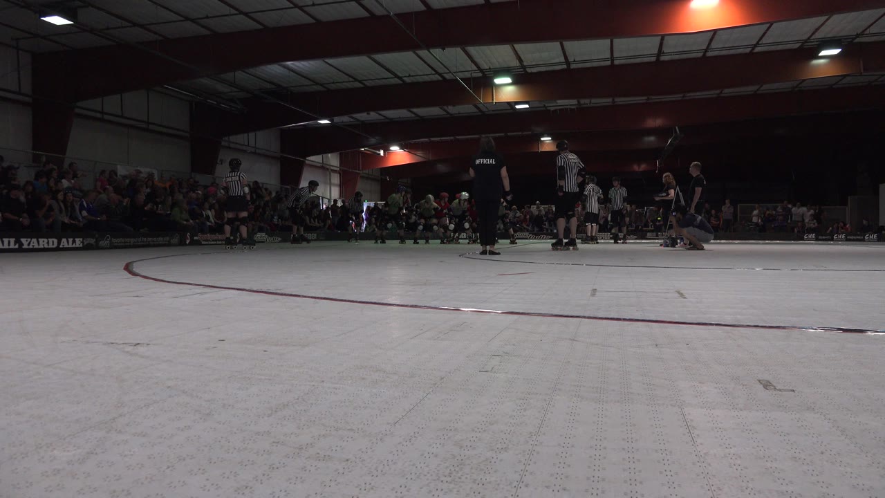 At the start of the Denver clip, a low camera looks across a pale indoor skating surface toward skaters, officials, and spectators gathered in the distance.