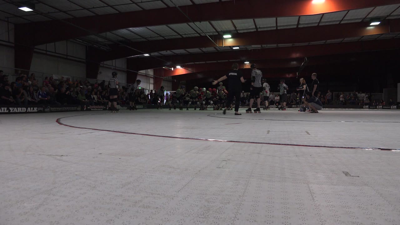 Low rink-level view in Denver, Colorado shows roller derby skaters and striped-shirt referees gathered near the starting area with spectators seated behind them.