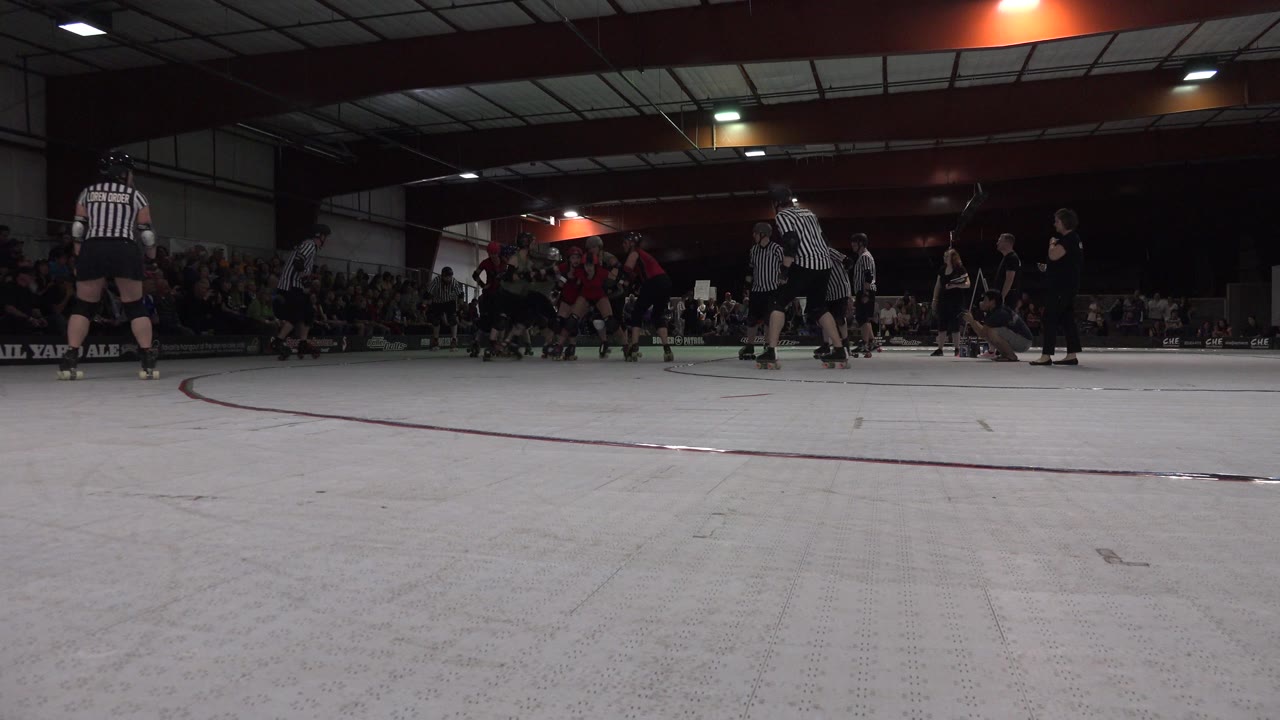Low court-level frame from Denver, Colorado showing roller skate players grouped near the left side, striped referees on the floor, and spectators seated in the dim background.