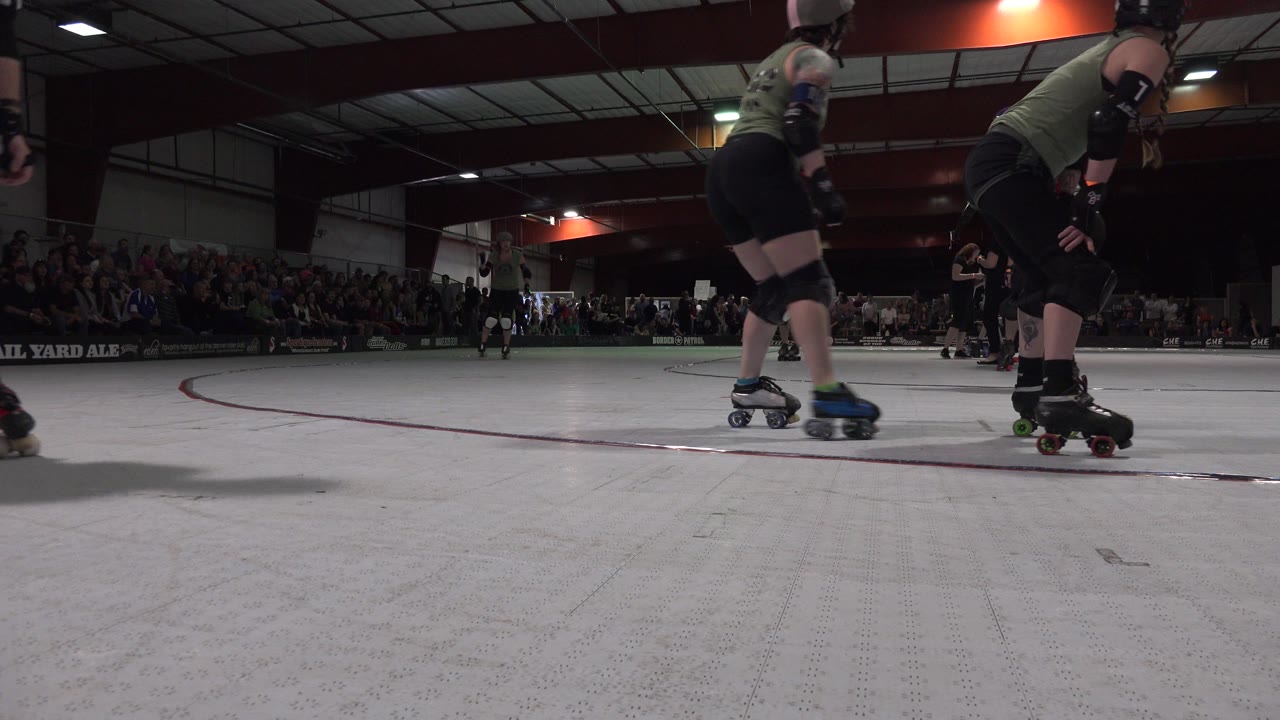 Denver indoor rink scene with two skaters in helmets and pads near the red floor line while the audience sits in shadow behind the track.