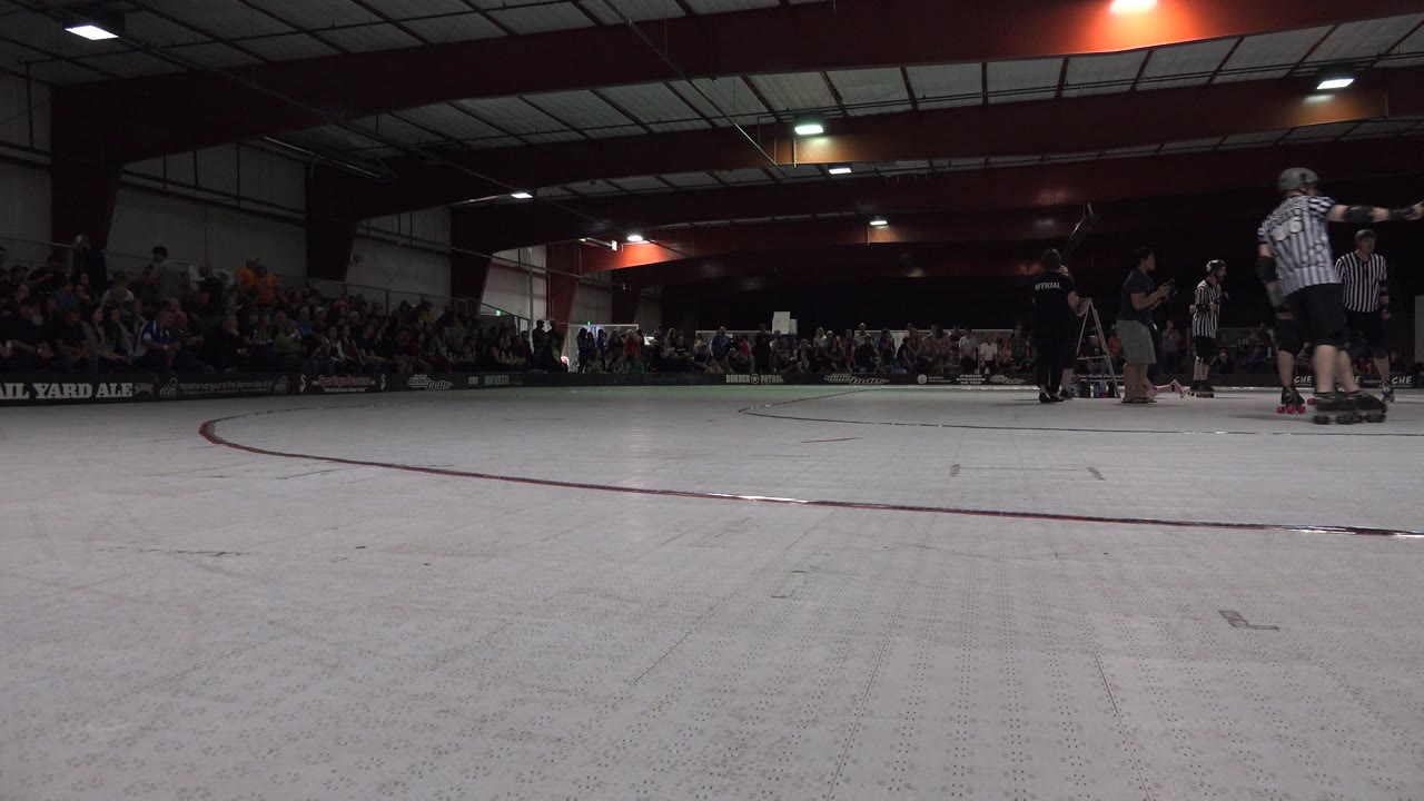 Low view of an indoor roller rink with skaters, officials, and spectators.