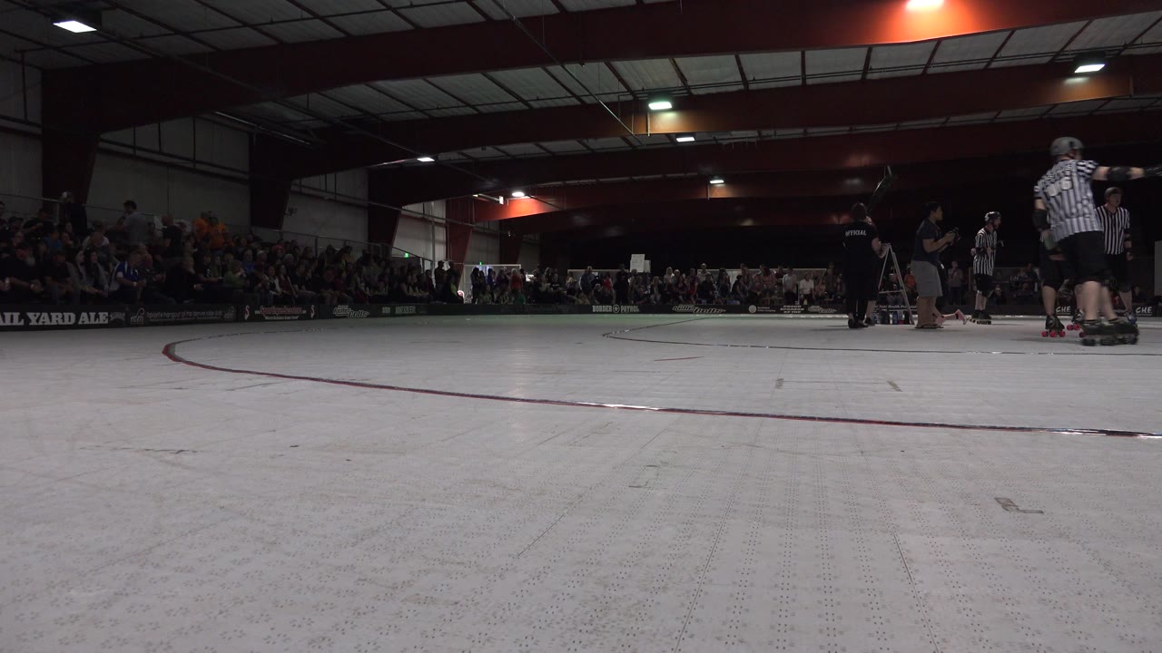 The Denver roller rink remains framed from floor level, with the broad pale surface dominating the foreground and several skaters standing near striped officials on the right.