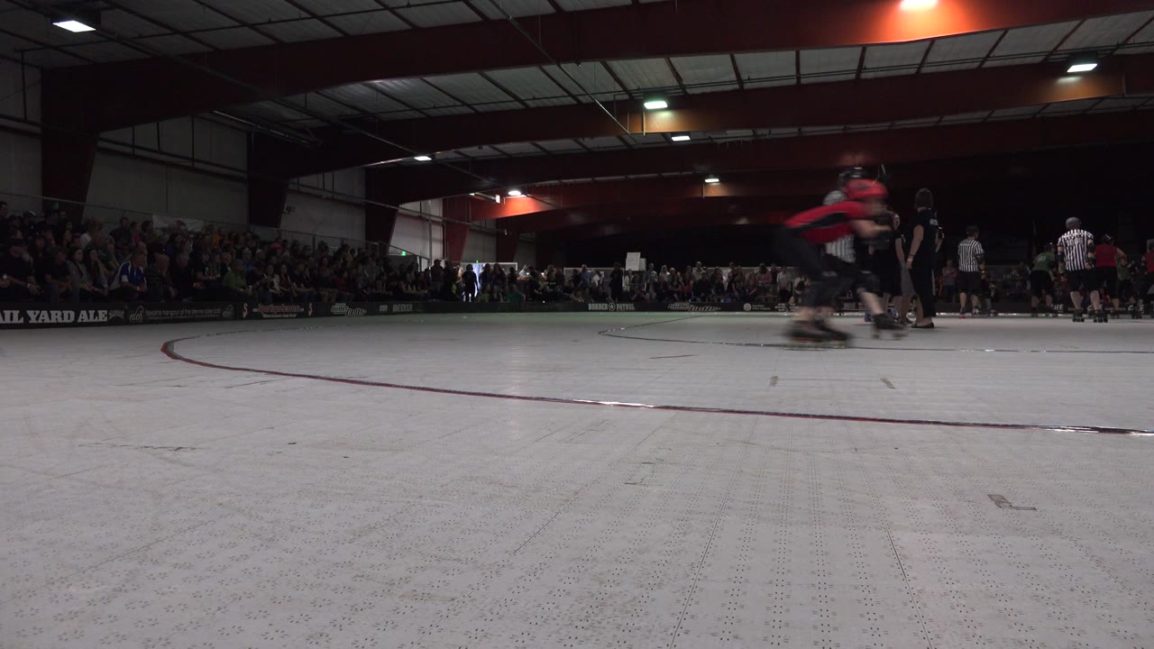 A skater in red moves across the Denver rink near the camera while other skaters and spectators remain in the dim background.