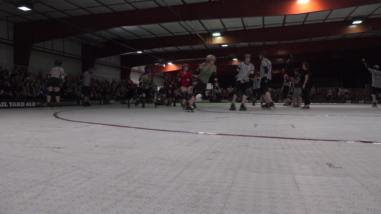 Denver indoor skating scene at rink level, with helmeted skaters in red and green gear, striped referees, and a seated audience behind the track.