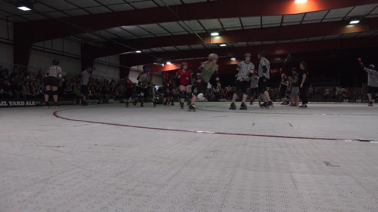 A low Denver rink view shows skaters moving through the midground while referees stand near the track and spectators watch from the dark back wall.