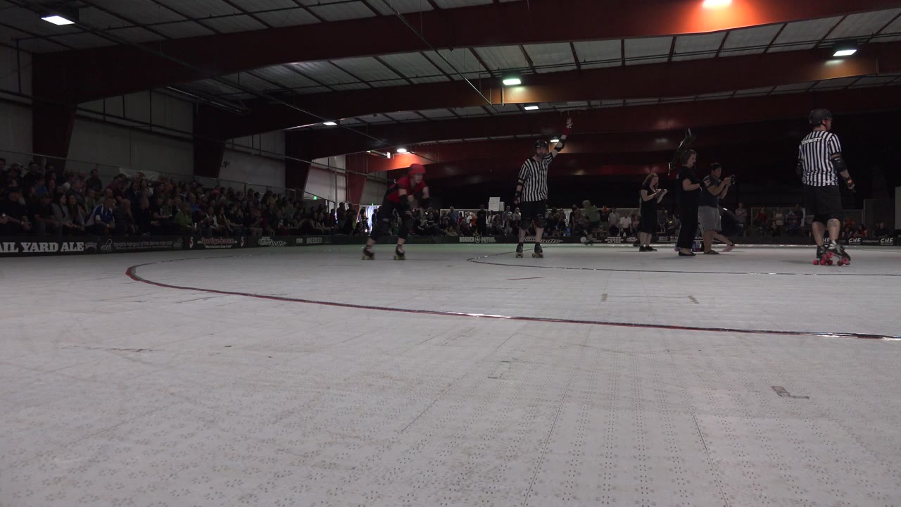 Low view of an indoor rollerblading floor with skaters, striped officials, and a seated crowd.