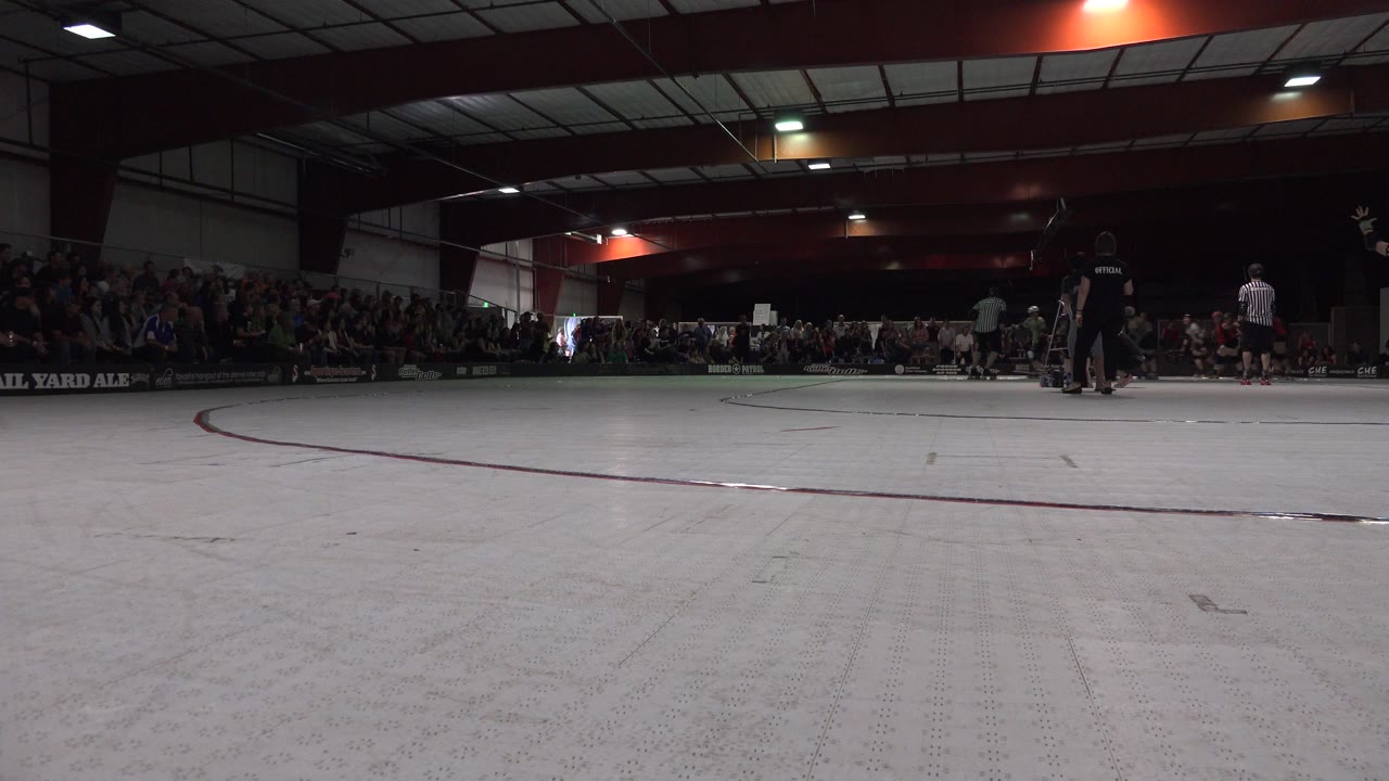Wide low view of roller derby skaters on an indoor track with spectators behind them.