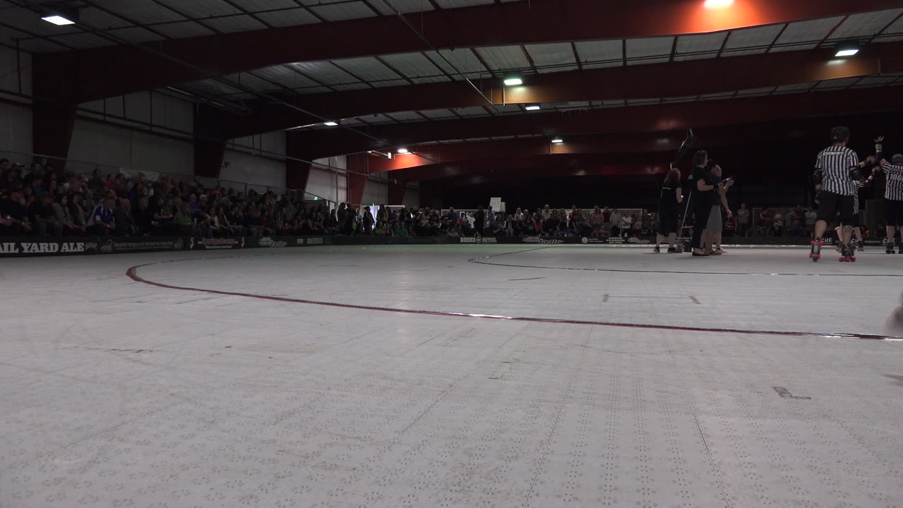 Denver indoor rink frame with open white floor in front, a small group of skaters and referees at mid-right, and a packed spectator area in the background.