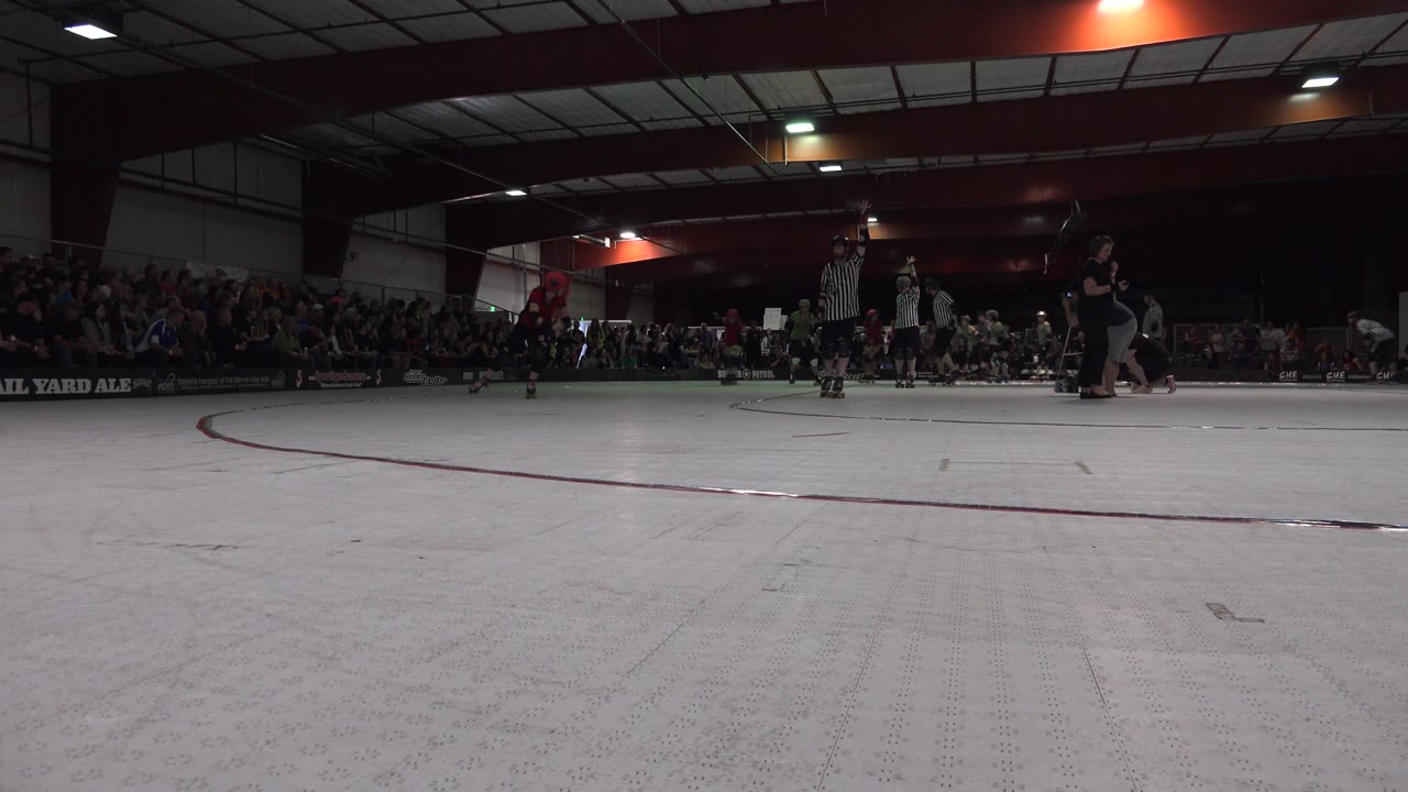 Low rink-side frame in Denver showing a pale skating floor, a red curved line, distant helmeted skaters, referees, and spectators along the back wall.