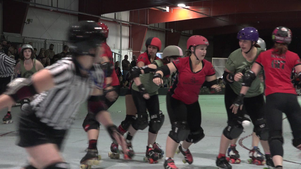 Denver roller derby players in helmets and pads skate close together while spectators sit behind the rink barrier under indoor venue lights.