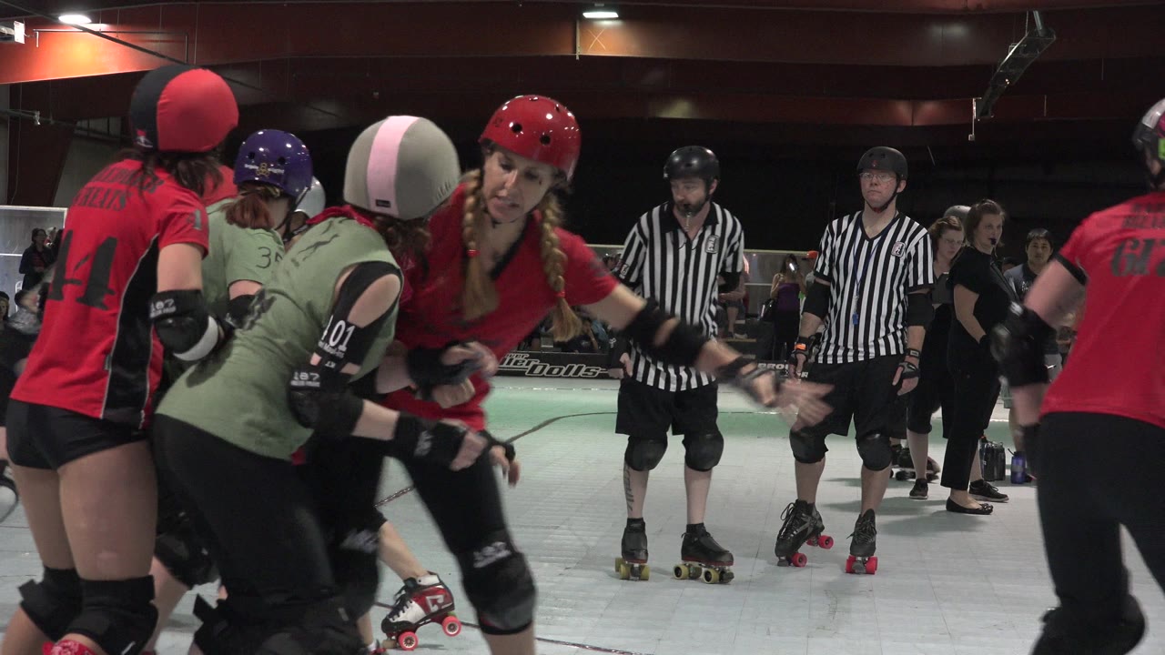 Roller derby skaters in red and green jerseys crowd together on an indoor track.