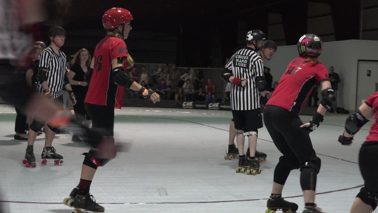 Denver skaters in red and black uniforms move near two referees on the indoor court while spectators watch from behind a barrier.