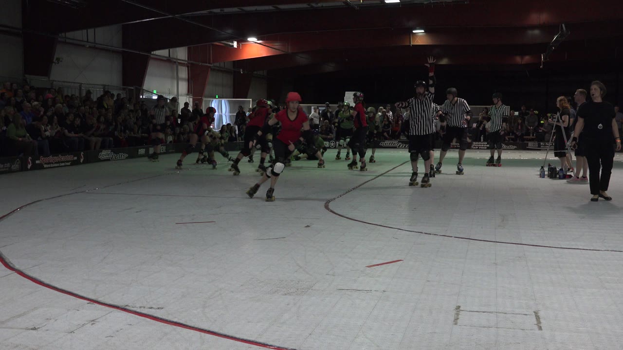 Denver indoor roller derby scene with red-uniformed women skaters approaching mid-rink while striped referees and a seated crowd are visible in the background.