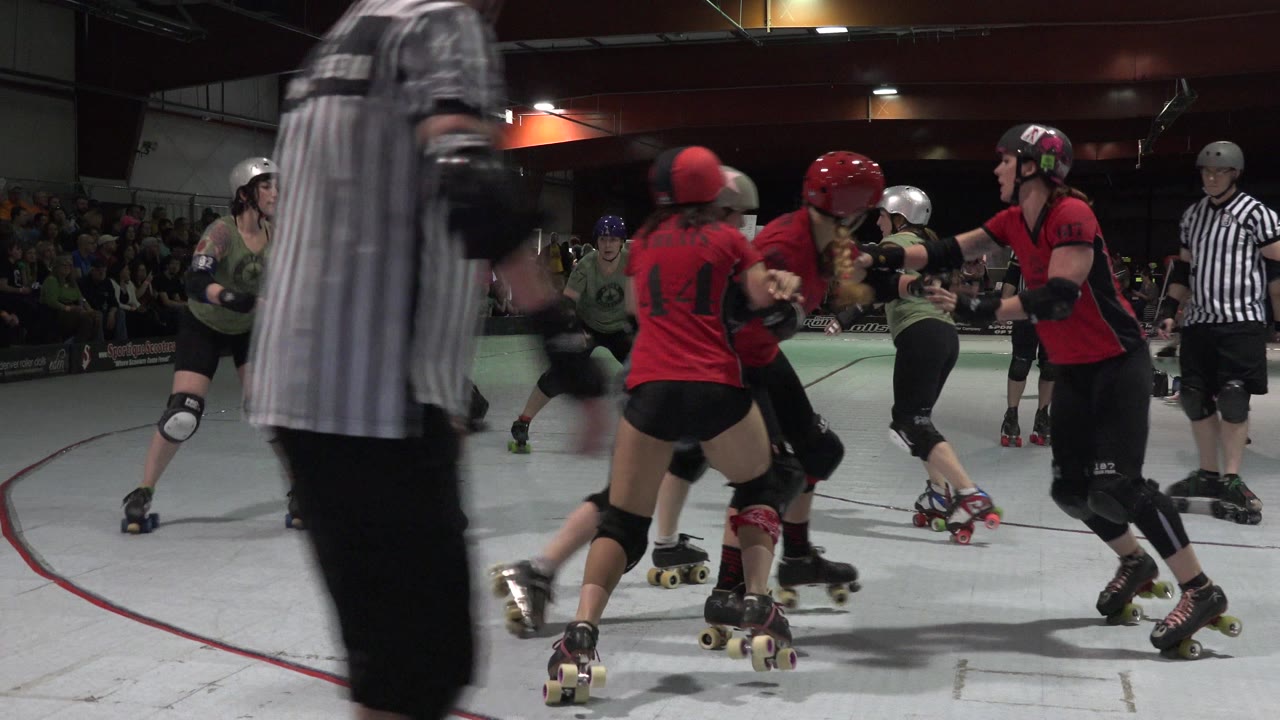 Roller derby skaters bunch on an indoor track as a referee crosses the foreground.