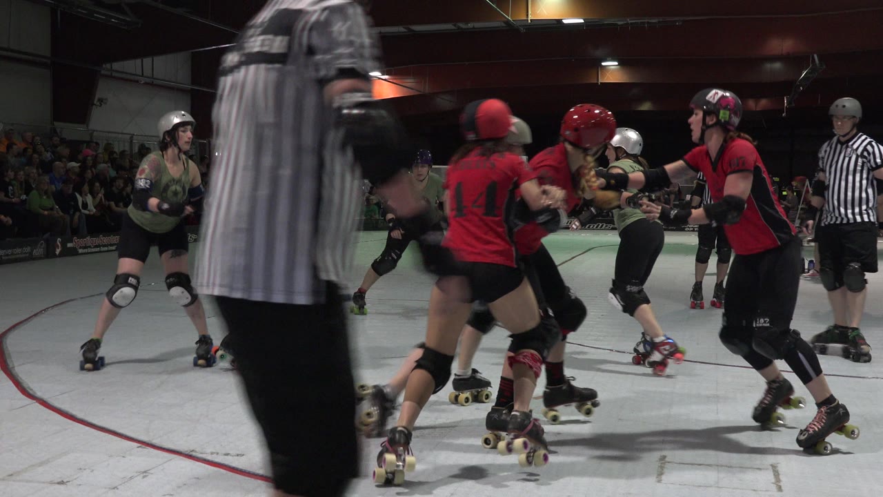 In Denver, Colorado, the roller derby pack stays tight as red-jersey and olive-jersey skaters lean into contact with a referee standing at the right edge.