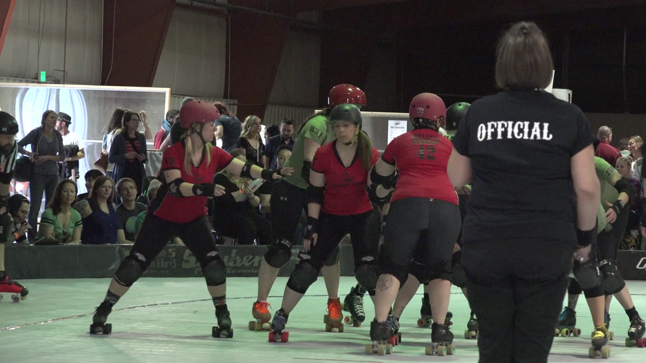 In Denver, Colorado, the skaters remain bunched together on the indoor rink while the official’s back fills the foreground and seated spectators look on from the background.