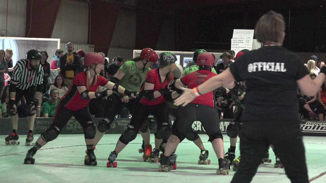 In Denver, Colorado, players wearing helmets and pads lean into each other on quad skates as a striped referee and seated spectators remain visible in the background.