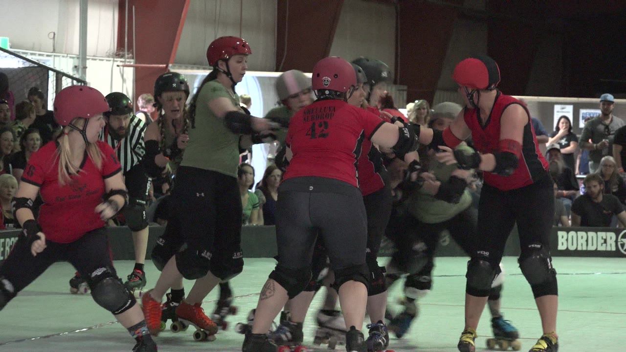 Denver, Colorado roller derby skaters in red and green jerseys bunch together on a pale green indoor track with spectators behind the barrier.
