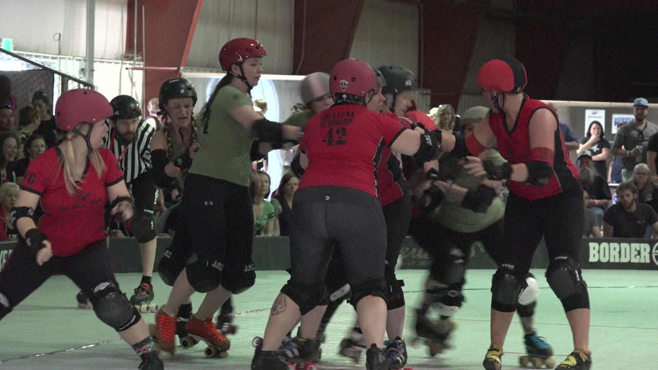 Denver, Colorado skaters in red and green roller derby uniforms push through a tight cluster as seated and standing spectators watch from behind the rink barrier.