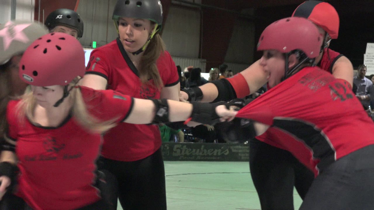 Helmeted women in red shirts crowd together during an indoor sports game.