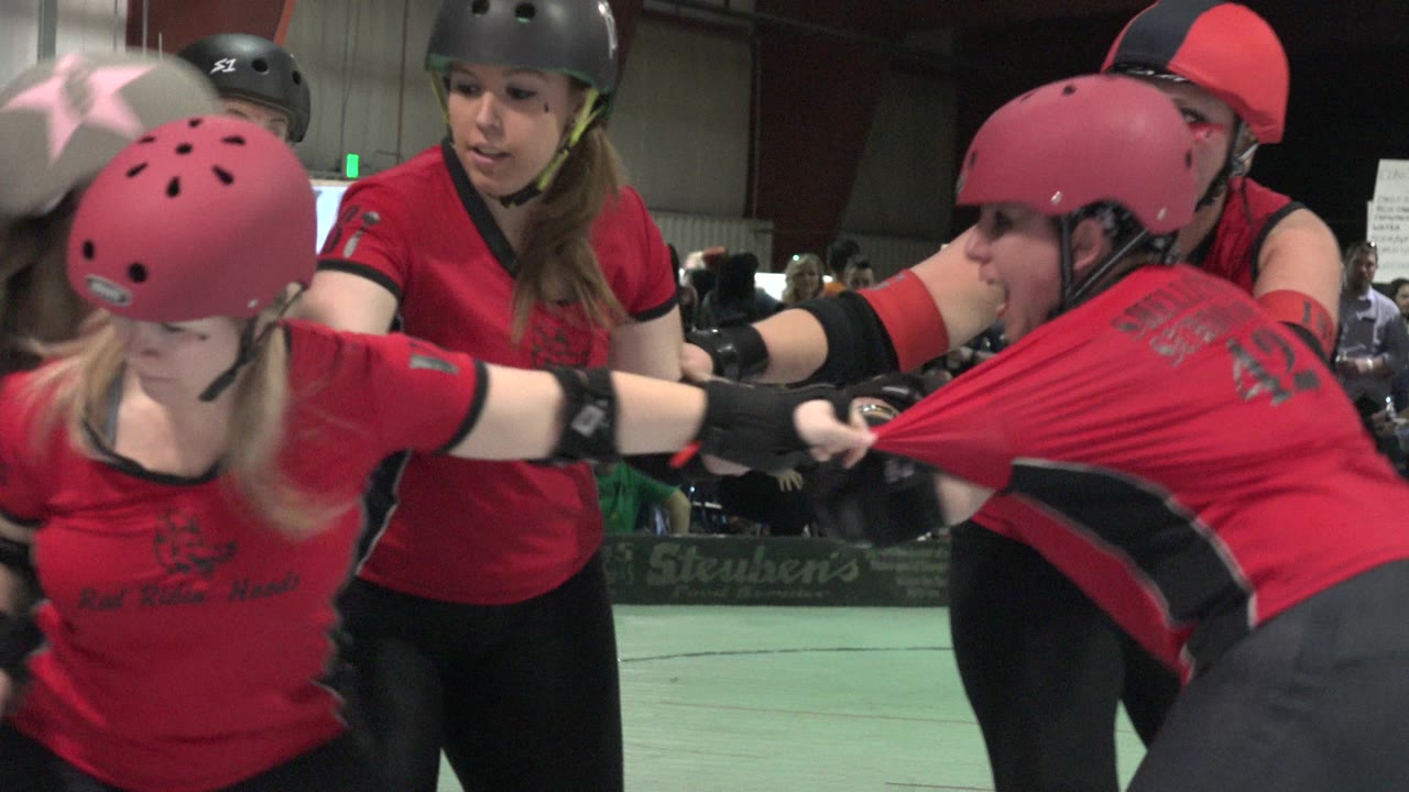 In Denver, Colorado, two red-shirted players in helmets lean into a tight foreground exchange while other athletes and spectators sit deeper in the indoor venue.