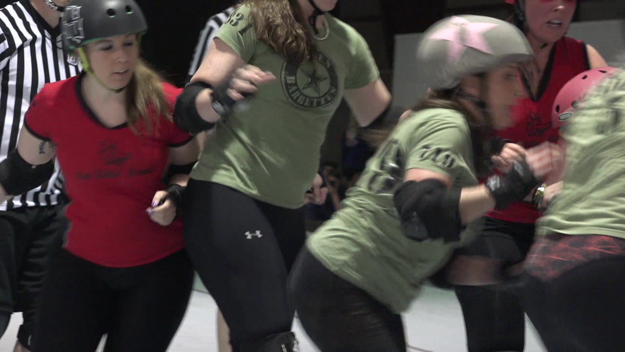 Roller derby skaters in red and green tops cluster near a striped referee.