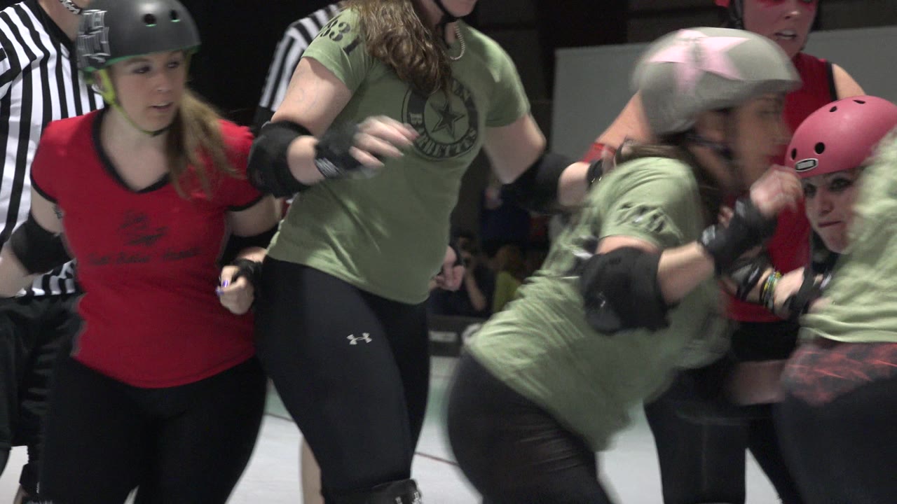 Denver rink action with a green-shirted skater leaning forward through traffic while red-team skaters, helmets, pads, and a referee fill the close foreground.
