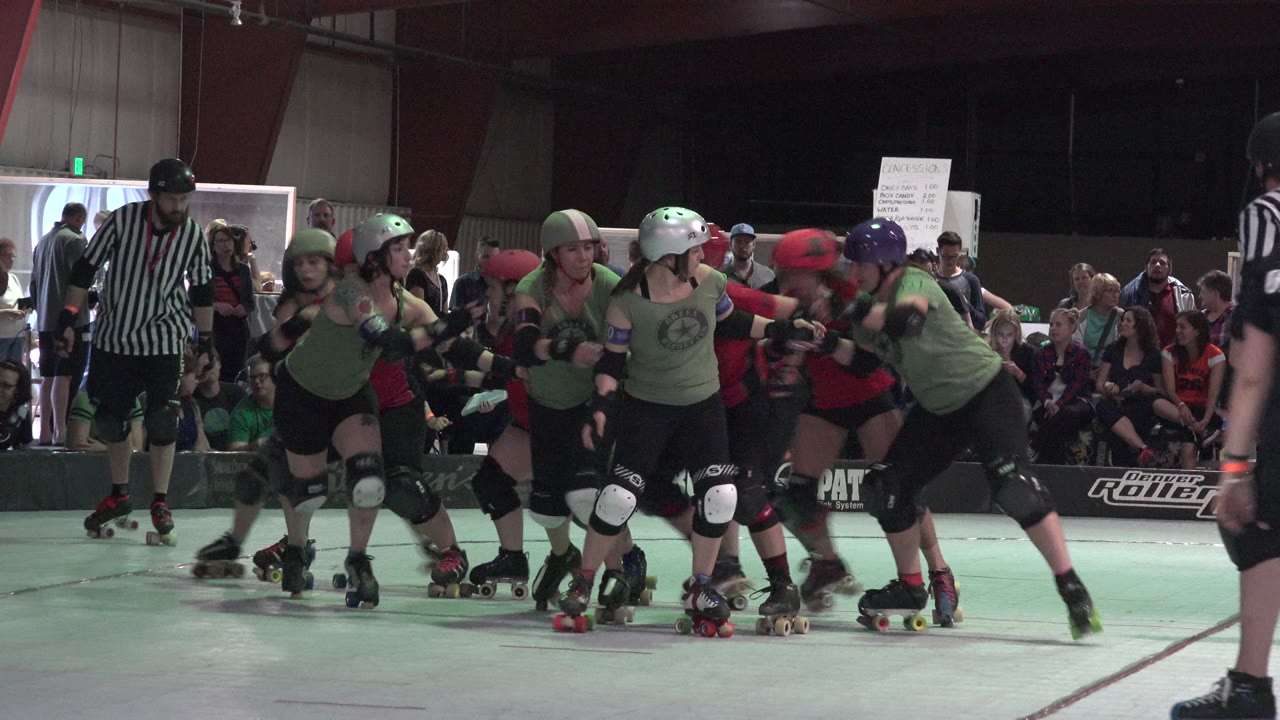 Denver 2015 roller derby skaters in protective gear press together on the track while a referee watches from the left and spectators fill the background.