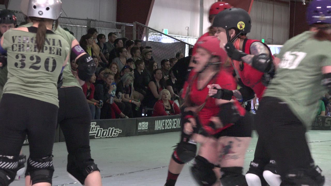 A red jersey skater is boxed in by green jersey skaters on the Denver indoor rink, with seated spectators watching from behind the boards.