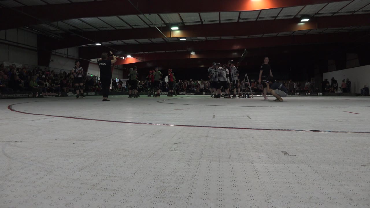Low gym floor view with skaters, referees, and spectators on bleachers.