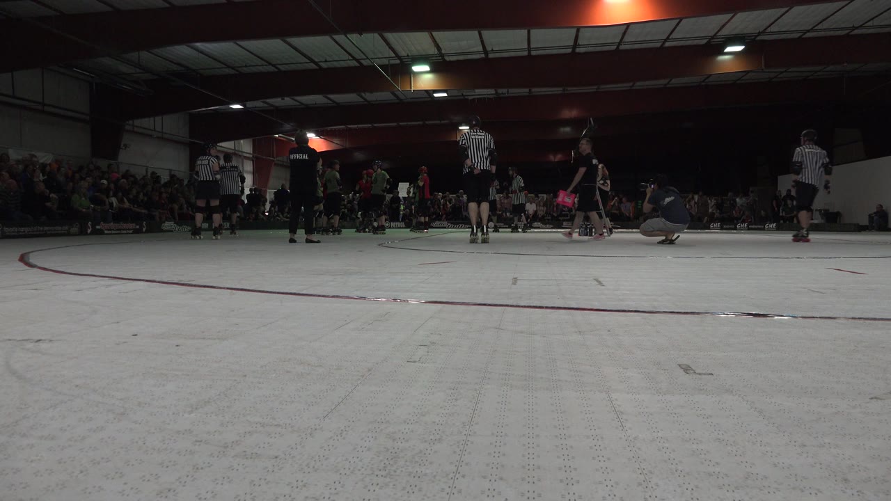 Low rink-level frame from Denver, Colorado showing teen roller players and striped referees in a dim gym with spectators along the back wall.