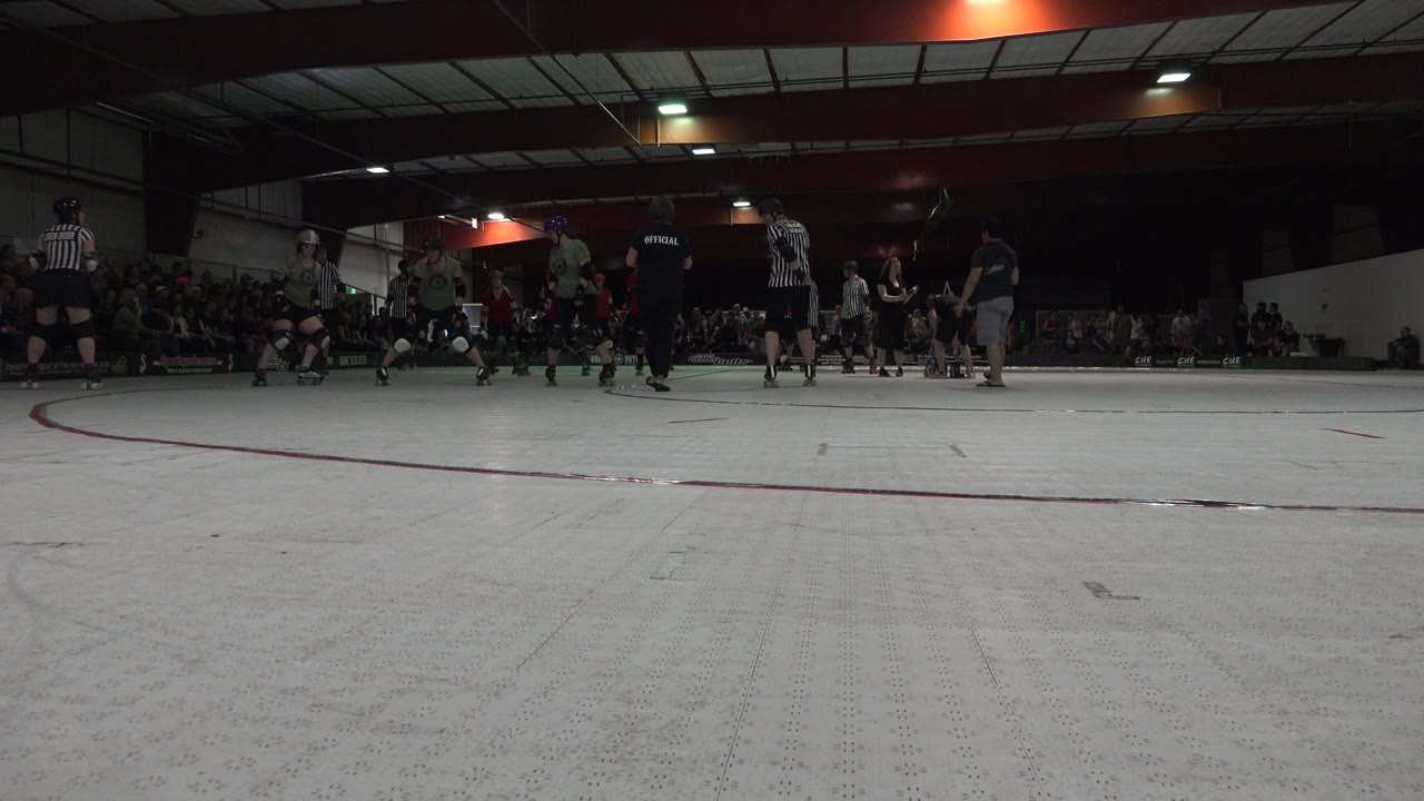 Low ground-level view in Denver, Colorado shows skaters and striped officials gathered on a white indoor roller rink with spectators seated in the background.