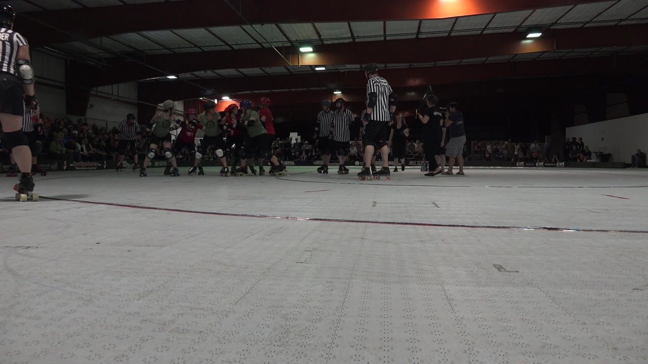 A low camera angle in Denver, Colorado frames the roller sport pack slightly farther across the rink, with a referee near center and spectators seated behind the track.